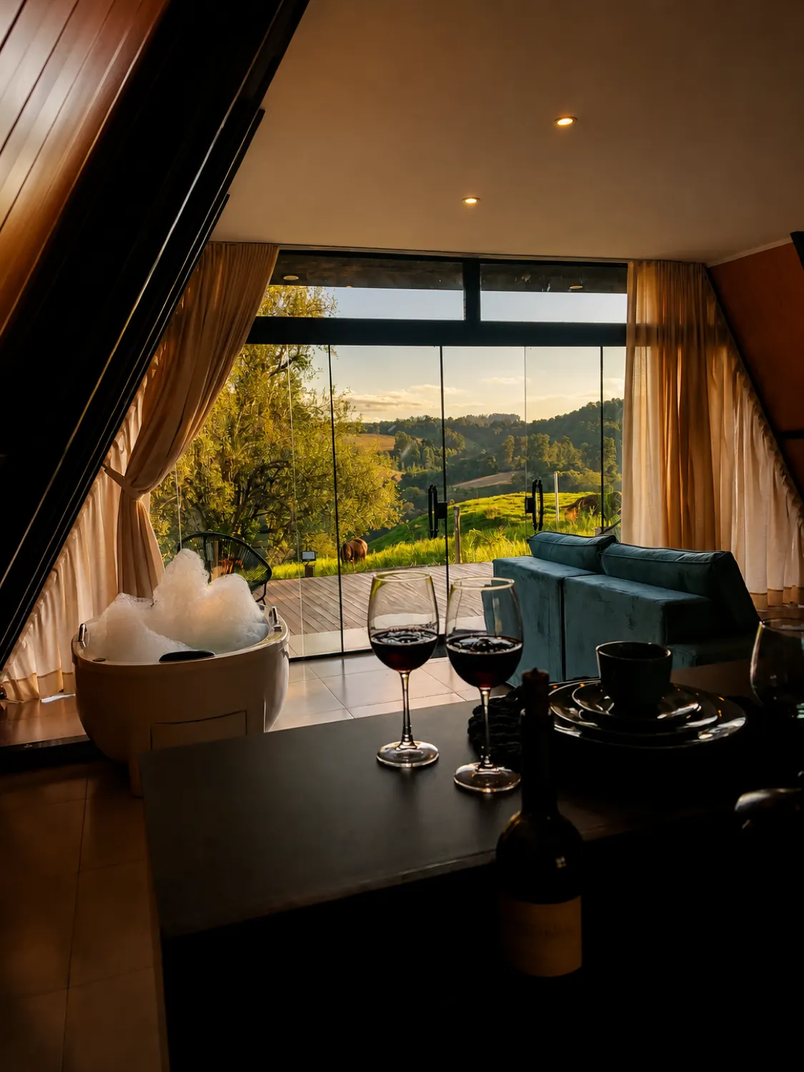 Sala do Chalé da Lagoa ao pôr do sol: sofá, taças de vinho e o vidro do chão ao teto com vista para o vale